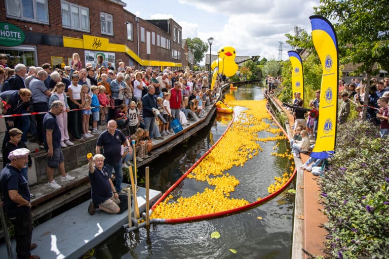 Publiek langs de kant kijkt toe terwijl duizenden badeendjes door de Hennipsloot dobberen